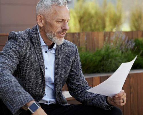 a man in grey blazer sitting on a bench and looking the piece of paper